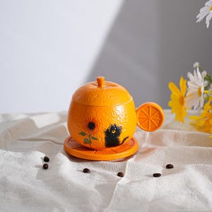May include: Orange-shaped ceramic mug with a lid and saucer. The mug features a black cat illustration and a sunflower design. The handle is shaped like an orange slice. The set is orange and sits on a white cloth.