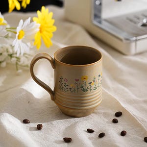 Puede incluir: Una taza de cer&aacute;mica beige con un dise&ntilde;o floral, con flores amarillas, rosas y azules. La taza tiene un asa curva y est&aacute; colocada sobre una tela blanca, con granos de caf&eacute; dispersos. Una cafetera se encuentra en el fondo.
