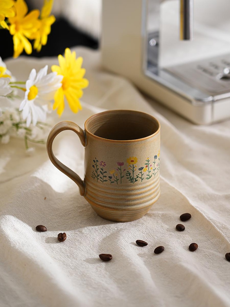 Puede incluir: Una taza de cer&aacute;mica beige con un dise&ntilde;o floral, con flores amarillas, rosas y azules. La taza tiene un asa curva y est&aacute; colocada sobre una tela blanca, con granos de caf&eacute; dispersos. Una cafetera se encuentra en el fondo.