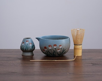 May include: A ceramic matcha tea set featuring a blue bowl with a spout, a small vase, a bamboo whisk, and a bamboo scoop. The set is decorated with a floral pattern in red, white, and green.