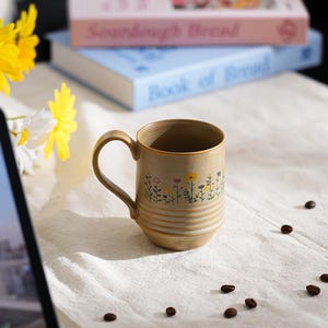 Puede incluir: Una taza de cer&aacute;mica beige con un dise&ntilde;o floral, colocada sobre una tela blanca. La taza tiene un asa curva y est&aacute; decorada con peque&ntilde;as flores de colores. Granos de caf&eacute; est&aacute;n esparcidos alrededor de la taza. Dos libros apilados en el fondo.