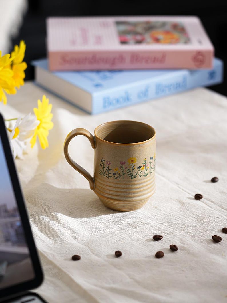 Puede incluir: Una taza de cer&aacute;mica beige con un dise&ntilde;o floral, colocada sobre una tela blanca. La taza tiene un asa curva y est&aacute; decorada con peque&ntilde;as flores de colores. Granos de caf&eacute; est&aacute;n esparcidos alrededor de la taza. Dos libros apilados en el fondo.