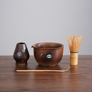 Może przedstawiać: Brązowy ceramiczny zestaw do herbaty matcha z miską, trzepaczką i małym wazonem. Miska i wazon mają wzór czaszki Grim Reaper. W zestawie znajduje się również bambusowa trzepaczka i miarka. Zestaw jest prezentowany na drewnianej powierzchni.
