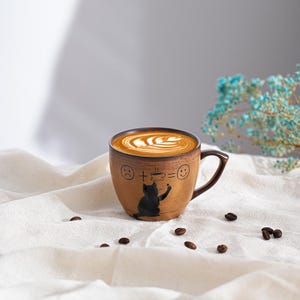 May include: A brown ceramic coffee mug with a dark brown handle and rim, filled with latte art. The mug features a black cat illustration and the equation: sad face + coffee = happy face. Coffee beans are scattered around the mug.