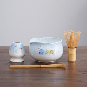 Puede incluir: Un juego de té matcha con un cuenco de cerámica azul claro con un diseño de animales de dibujos animados, un batidor, un pequeño recipiente de cerámica y una cuchara de bambú. El juego está dispuesto sobre una superficie de madera.