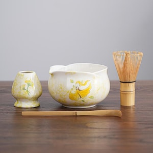 以下が含まれることがあります： 白いセラミックボウル、黄色の花柄、竹の泡立て器、小さなセラミックホルダー、竹のスクープを備えた抹茶ティーセット。 セットは木製の表面に配置されています。