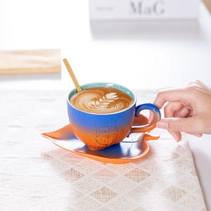 May include: A blue and orange gradient ceramic coffee cup with a cat design, filled with latte art, sits on a matching leaf-shaped saucer. A small wooden spoon rests in the cup. The cup is held by a person's hand. The background includes a white surface and a patterned tablecloth.