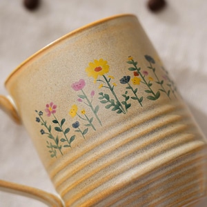 Puede incluir: Una taza de cer&aacute;mica beige con un dise&ntilde;o floral. La taza tiene un asa y estr&iacute;as horizontales. El dise&ntilde;o floral incluye flores amarillas, rosas y azules con tallos y hojas verdes. La taza es adecuada para bebidas calientes.