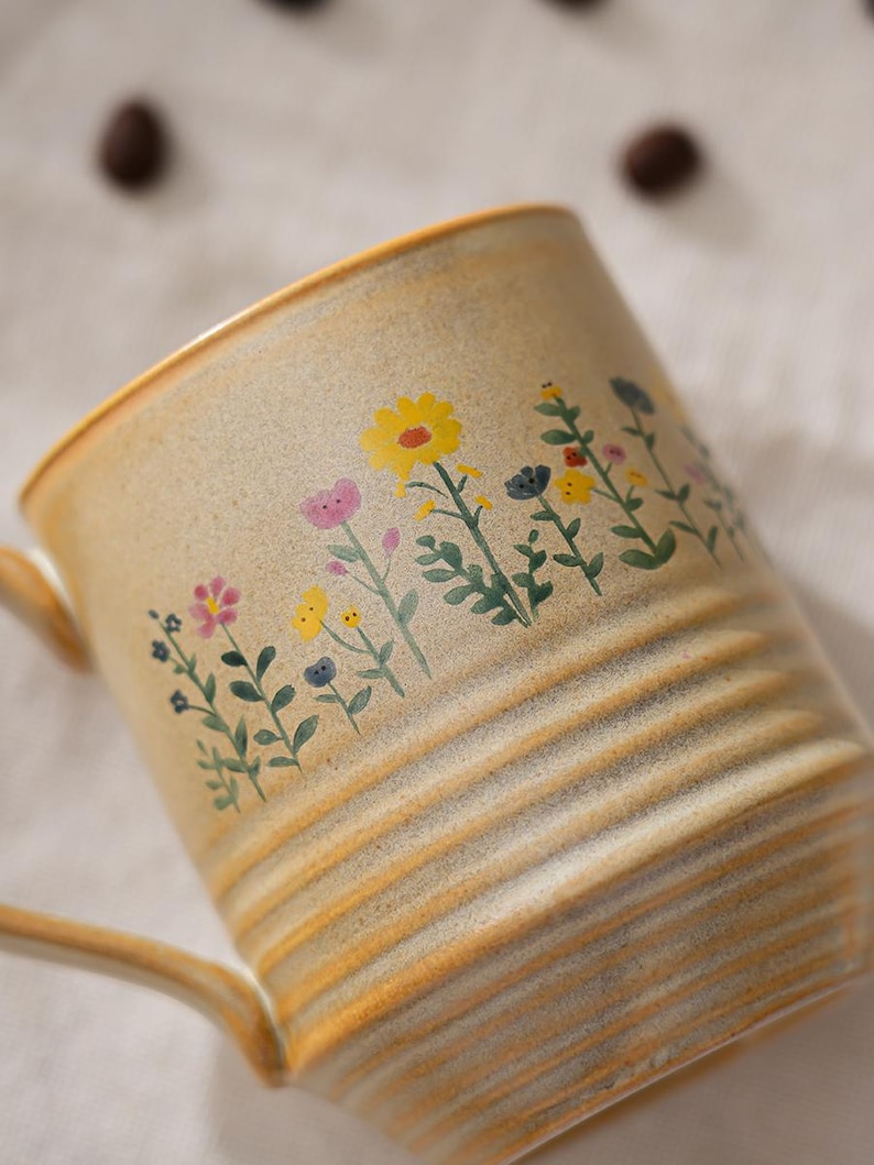 Puede incluir: Una taza de cer&aacute;mica beige con un dise&ntilde;o floral. La taza tiene un asa y estr&iacute;as horizontales. El dise&ntilde;o floral incluye flores amarillas, rosas y azules con tallos y hojas verdes. La taza es adecuada para bebidas calientes.