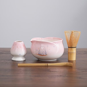 Puede incluir: Un juego de té matcha de cerámica rosa con un diseño de fantasma. El juego incluye un cuenco, un batidor, un soporte para batidor y una cuchara. El cuenco y el soporte tienen un esquema de color rosa y blanco degradado. El batidor es de bambú.