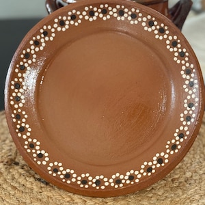 May include: A brown ceramic plate with a decorative border of white and black dots. A matching brown ceramic pot is visible in the background. The plate is placed on a woven mat. The plate is part of a set.