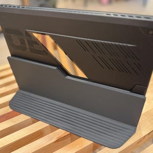 Puede incluir: Un ordenador portátil gris oscuro con un diseño único, con un soporte y una parte trasera estampada. El portátil está en ángulo, mostrando su perfil lateral con una ventilación y un recorte geométrico. El soporte tiene una textura acanalada.