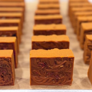 May include: Close-up of several handmade, rectangular soaps in a warm orange colour. The soaps have a swirled pattern and botanical inclusions. The soaps are arranged in rows on a light-coloured surface.