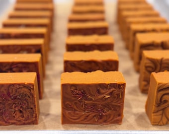 May include: Close-up of several handmade, rectangular soaps in a warm orange colour. The soaps have a swirled pattern and botanical inclusions. The soaps are arranged in rows on a light-coloured surface.