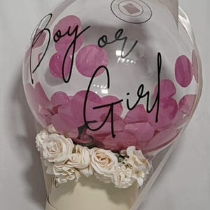 May include: A clear balloon filled with pink confetti, featuring the black text "Boy or Girl" printed on the surface. The balloon is attached to a cream-coloured cylindrical base decorated with a ring of white artificial roses and a white ribbon.