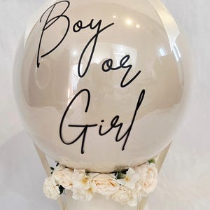 May include: A large, round, beige balloon featuring the black cursive text "Boy or Girl" on its surface. The balloon is topped with a cream-coloured ribbon bow and is decorated at its base with a ring of cream-coloured artificial roses.