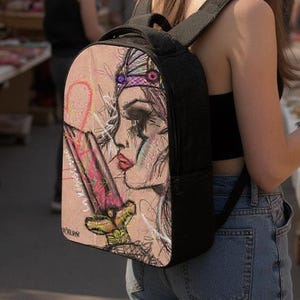 Puede incluir: Mochila negra con una colorida ilustración de una mujer con una espada. La obra de arte presenta tonos de rosa, rojo y dorado. La mochila la lleva una persona con un top negro y vaqueros azules, que está mirando un mercado al aire libre.