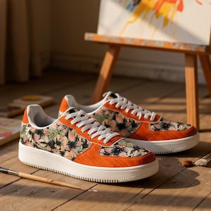 May include: A pair of orange and floral print sneakers with white soles and laces. The sneakers are on a wooden floor with paintbrushes and a painting in the background. The sneakers have a low-top design.
