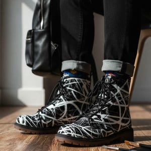 Puede incluir: Botas de combate en blanco y negro con un diseño abstracto. Las botas tienen cierre de cordones, punta redonda y suela marrón. Las botas se llevan con vaqueros negros. Una mochila negra es visible en el fondo. Las botas miden unos 25 cm de largo.