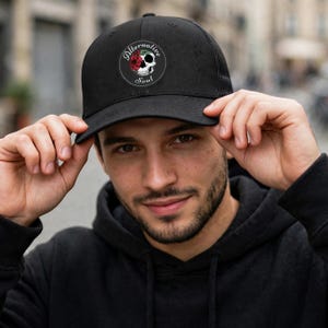 Peut inclure: Une casquette de type trucker noire avec un écusson circulaire sur le devant. L'écusson présente un crâne blanc orné d'une rose rouge et de feuilles vertes, entouré du texte Alternative Soul en police cursive blanche sur fond noir.