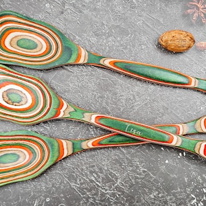 May include: Three wooden spoons with a green, orange, and white swirl design. One spoon has the name "Lisa" engraved on the handle. The spoons have a hole at the end of the handle for hanging.