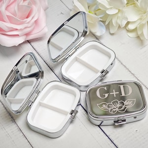 May include: Silver pill boxes with white interiors and mirrors. One box is closed, engraved with the initials "G+D" and a floral design. The boxes are open, revealing divided compartments. A pink rose and white flowers are in the background.