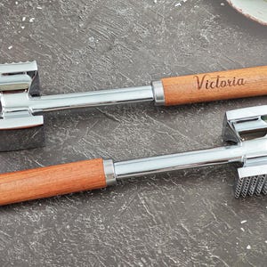 May include: Two stainless steel meat tenderizers with wooden handles. One handle is engraved with the name "Victoria". The tenderizers have a flat side and a textured side for different uses. The handles have a hole for hanging.