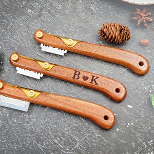 Op de afbeelding: Drie houten broodmessen met gouden accenten en vervangbare messen. Eén gereedschap is gegraveerd met "B♥K". De gereedschappen zijn gerangschikt op een donkergrijze ondergrond, met een dennenappel en andere decoratieve elementen.