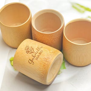 Può includere: Quattro tazze cilindriche di bambù. Una tazza è sdraiata su un lato, mostrando il testo inciso "Gabriella 16.8.2025" con un motivo floreale. Le altre tre tazze sono in posizione verticale, mostrando la venatura naturale del legno e gli interni aperti.