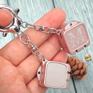 May include: A rose gold keychain with two square pendants connected by a chain. One pendant has the engraved name "Meechle" with a stylized initial "M". The keychain has a silver clasp and chain.