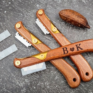 May include: Two wooden bread lame tools with gold accents and replaceable blades. One tool is engraved with "B ♥ K". Includes two blade covers and a decorative, textured, brown object. The tools are on a dark gray surface.