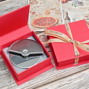 Może przedstawiać: Krajalnica do pizzy ze stali nierdzewnej w czerwonym pudełku prezentowym. Pudełko jest zawiązane wstążką z napisem "JUST FOR YOU". Krajalnica do pizzy znajduje się w otwartym czerwonym pudełku, a obok zamknięte czerwone pudełko.