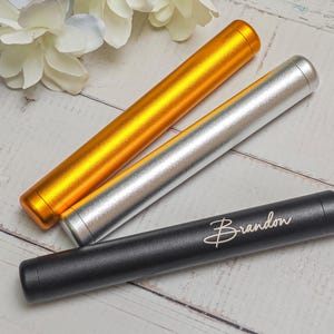 May include: Three cylindrical cases in gold, silver, and black. The black case has the name "Brandon" in cursive script. The cases are arranged on a light wood surface with white flowers in the background.