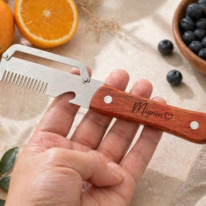 Puede incluir: Un pelador de frutas de acero inoxidable con mango de madera. El mango está grabado con la palabra "Mignon" y un símbolo de corazón. El pelador también tiene un borde en forma de peine y un abrebotellas.