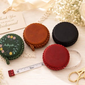 Peut inclure: Quatre mètres rubans ronds en cuir, vert, marron, noir et rouge. Un ruban à mesurer blanc est déployé. Une étiquette dit "Thank You For Being My Bridesmaid". Ciseaux dorés et éléments floraux complètent la scène.