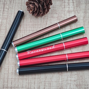 Puede incluir: Un surtido de tubos metálicos cilíndricos de colores, incluyendo negro, bronce, verde y rojo, con detalles plateados. Un tubo rojo lleva la inscripción "Ravenswood". Los tubos están dispuestos sobre una superficie de madera marrón claro.