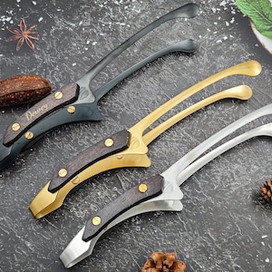 May include: Three metal tongs with wooden handles are displayed on a dark surface. One is black, one is gold, and one is silver. The wooden handles are dark brown and secured with brass-coloured rivets. The word "Dewey" is engraved on one of the handles.