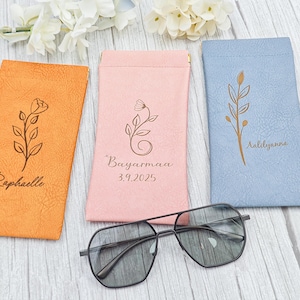 May include: An orange, pink, and blue glasses case set with a pair of black-framed sunglasses. Each case is personalized with a floral design and a name or date. The sunglasses have a geometric shape.