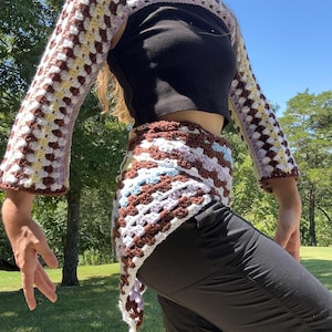 Puede incluir: Conjunto de bolero y falda envolvente de ganchillo, con patrón de cuadrados de la abuela. El bolero tiene mangas largas y un diseño corto, con marrón, blanco, lavanda y amarillo. La falda es de estilo envolvente, también en la misma combinación de colores.