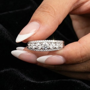 May include: A silver ring set with multiple round diamonds. The ring has a central row of larger diamonds, framed by smaller ones. The ring is displayed against a black backdrop.
