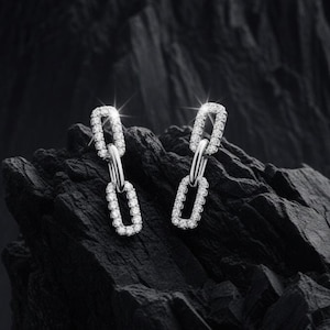 Puede incluir: Un par de pendientes plateados brillantes. Cada pendiente presenta un diseño de eslabones de cadena, con los eslabones incrustados con pequeñas piedras preciosas transparentes. Los pendientes se exhiben sobre un fondo oscuro y texturizado.