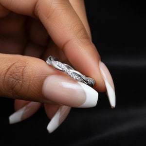 Puede incluir: Un anillo plateado con un diseño trenzado, adornado con pequeñas piedras preciosas transparentes. El anillo se sostiene sobre un fondo oscuro, mostrando sus intrincados detalles y brillo. El diseño del anillo es una trenza continua.