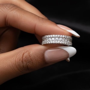 Puede incluir: Un anillo plateado con tres filas de pequeñas piedras preciosas transparentes. El anillo se sostiene entre los dedos sobre un fondo negro. Las piedras preciosas brillan, reflejando la luz.