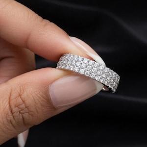 May include: A silver ring with three rows of small, clear gemstones. The ring is held between fingers against a black background, showcasing its sparkle and design. The ring is a piece of jewellery.