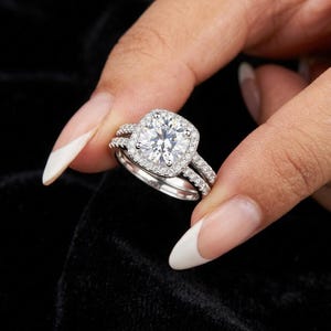 Peut inclure: Une bague en métal argenté avec une pierre centrale taille coussin entourée d'un halo de petites pierres rondes. L'anneau est orné d'une rangée de pierres serties pavé, avec une alliance assortie positionnée contre la bague principale.