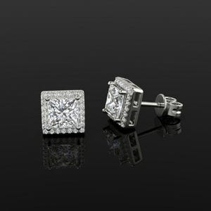 Może przedstawiać: Para kolczyków sztyftów z diamentami o kwadratowym szlifie z otoczką z mniejszych diamentów osadzonych w srebrnym metalu. Kolczyki są prezentowane na czarnej, odblaskowej powierzchni, podkreślając ich blask i design.