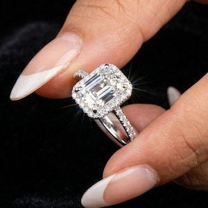May include: A sparkling, emerald-cut diamond ring with a halo of smaller diamonds. The ring features a silver band and is held against a dark background, showcasing its brilliance. A classic piece of jewelry.