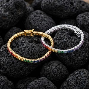 May include: Two tennis bracelets featuring a continuous row of multi-colored gemstones in a rainbow gradient. One bracelet is set in a gold-toned metal, while the other is set in a silver-toned metal, both featuring a secure box clasp closure.