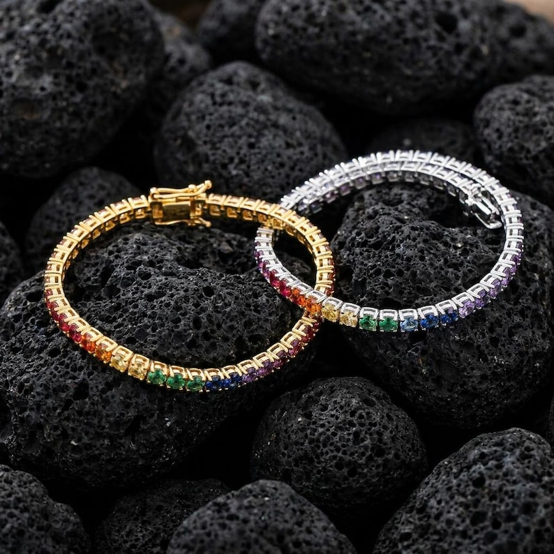 May include: Two tennis bracelets featuring a continuous row of multi-colored gemstones in a rainbow gradient. One bracelet is set in a gold-toned metal, while the other is set in a silver-toned metal, both featuring a secure box clasp closure.