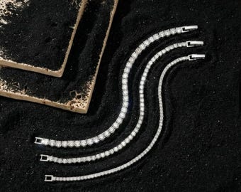May include: Three silver tennis bracelets with sparkling white gemstones are arranged on a black surface. The bracelets are of varying widths and have a classic, elegant design. A light-colored, textured rectangular object is in the upper left corner.